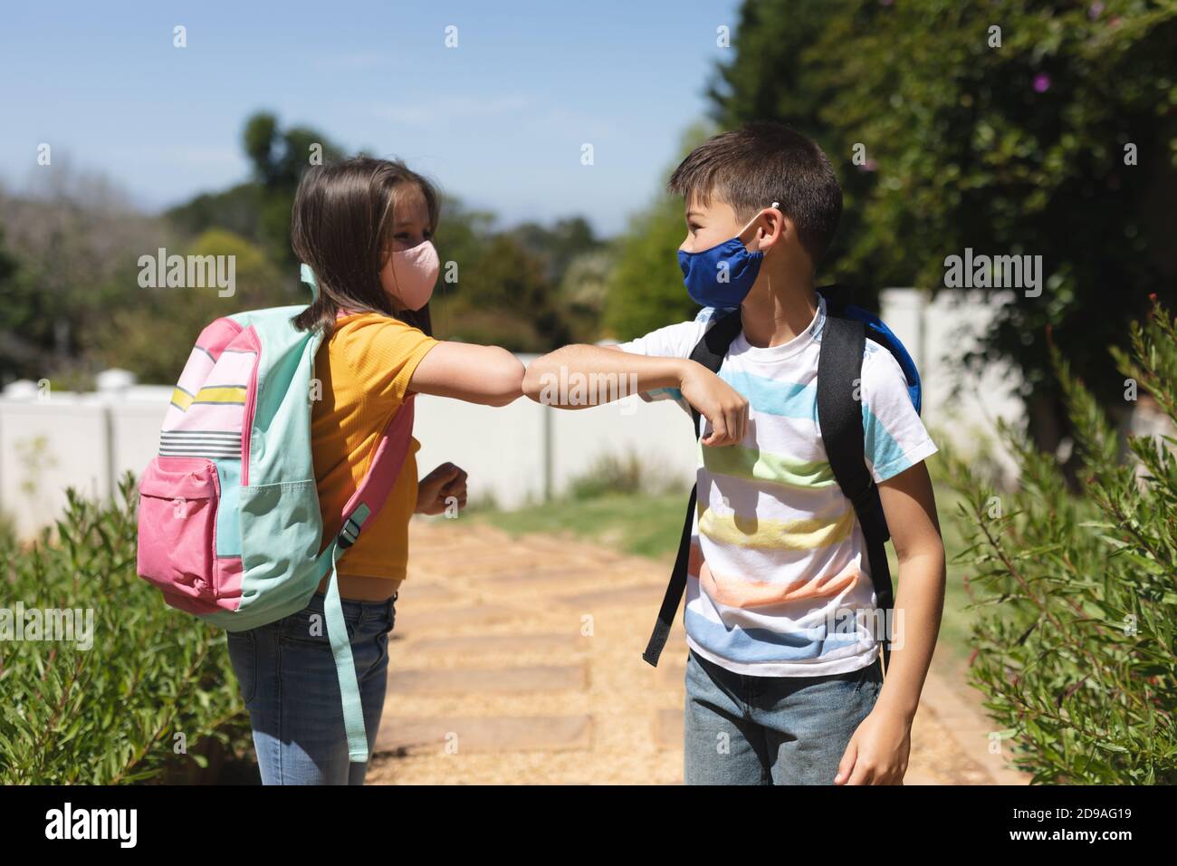 Ragazzo e ragazza caucasica che indossano maschere di saluto sul loro modo di scuola toccando gomiti Foto Stock