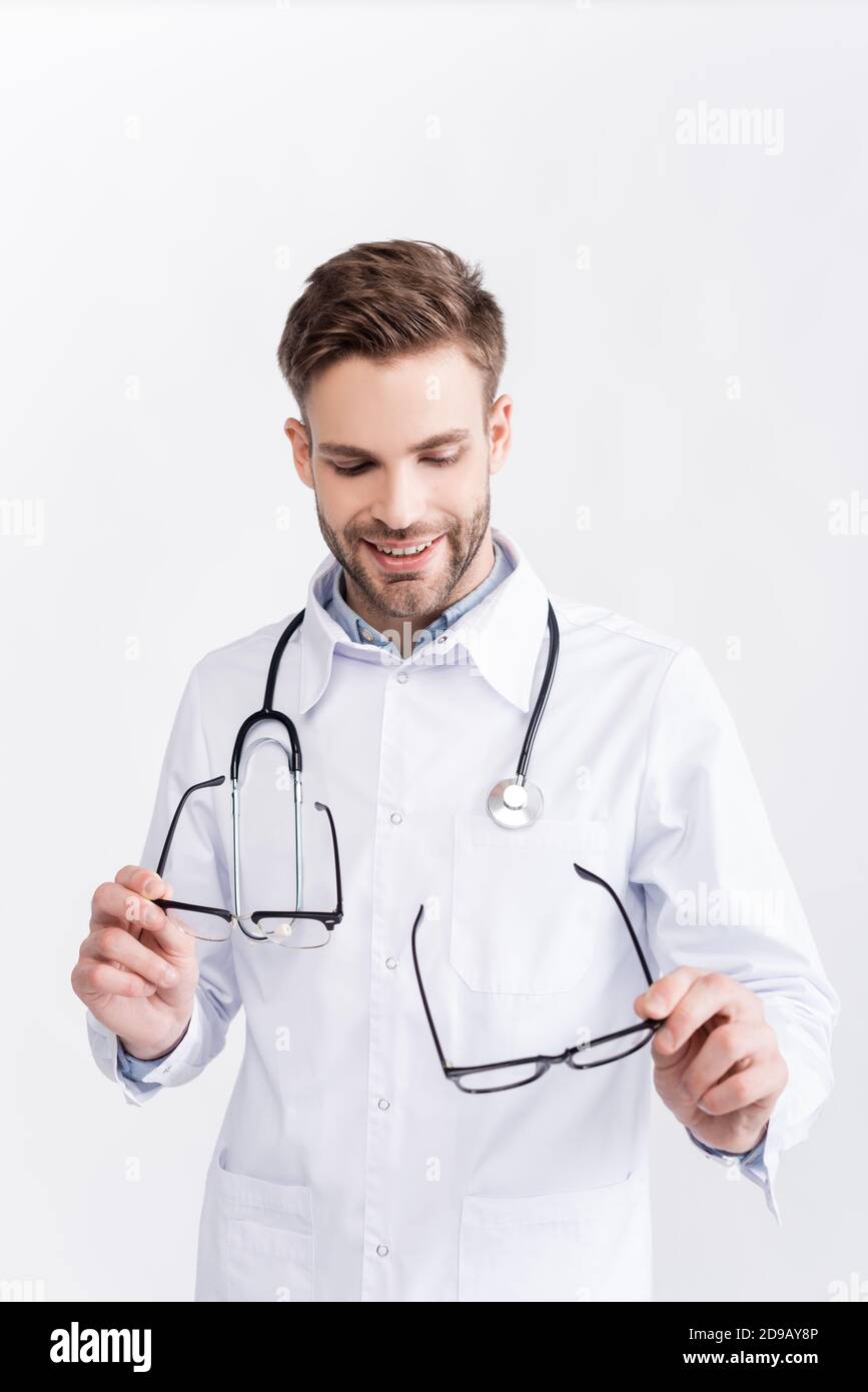 Sorridente medico che guarda e tiene un paio di occhiali con cornici nere isolate su bianco Foto Stock