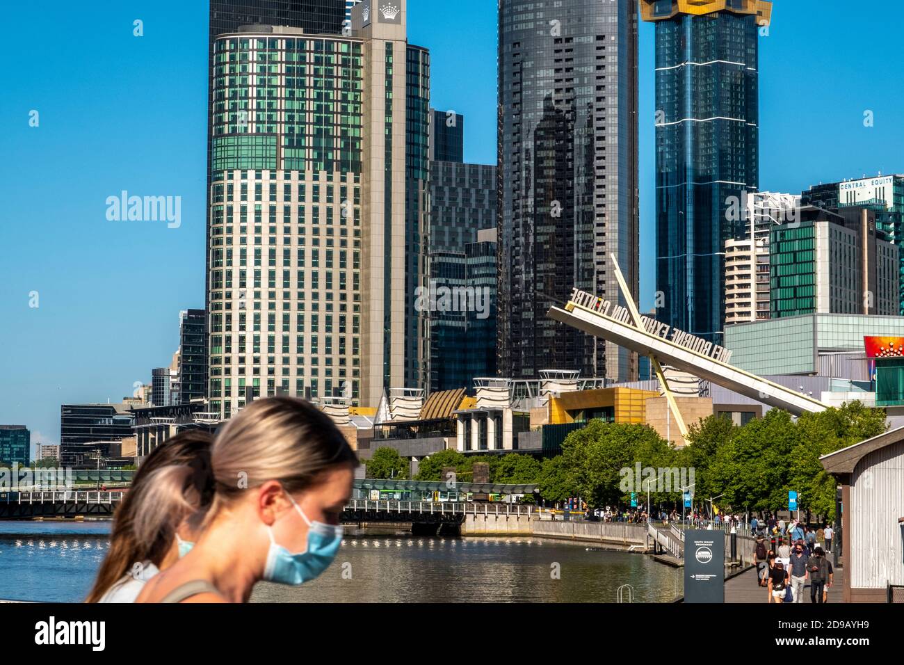 Persone mascherate camminano di fronte allo skyline della città di Melbourne. Foto Stock