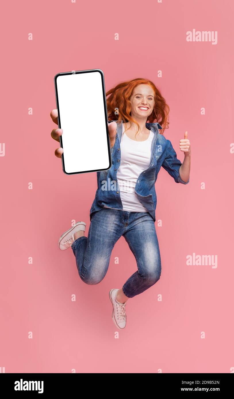 Ragazza redhead felice saltando con smartphone e mostrando il pollice in su Foto Stock