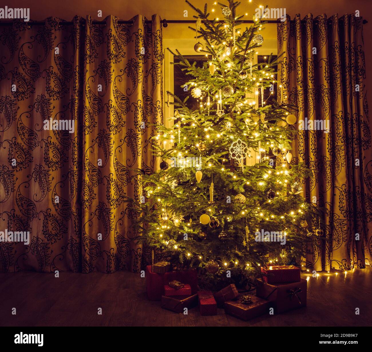 Albero di Natale con luci a corda calde gialle e sicli bianchi e ornamenti a forma di fiocco di neve. Regali rosso imballati sotto l'albero. Foto Stock