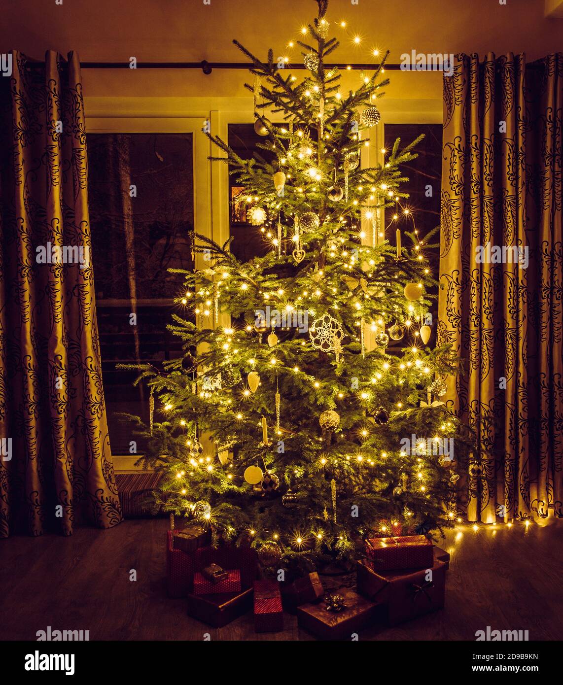 Albero di Natale con luci a corda calde gialle e sicli bianchi e ornamenti a forma di fiocco di neve. Regali rosso imballati sotto l'albero. Foto Stock
