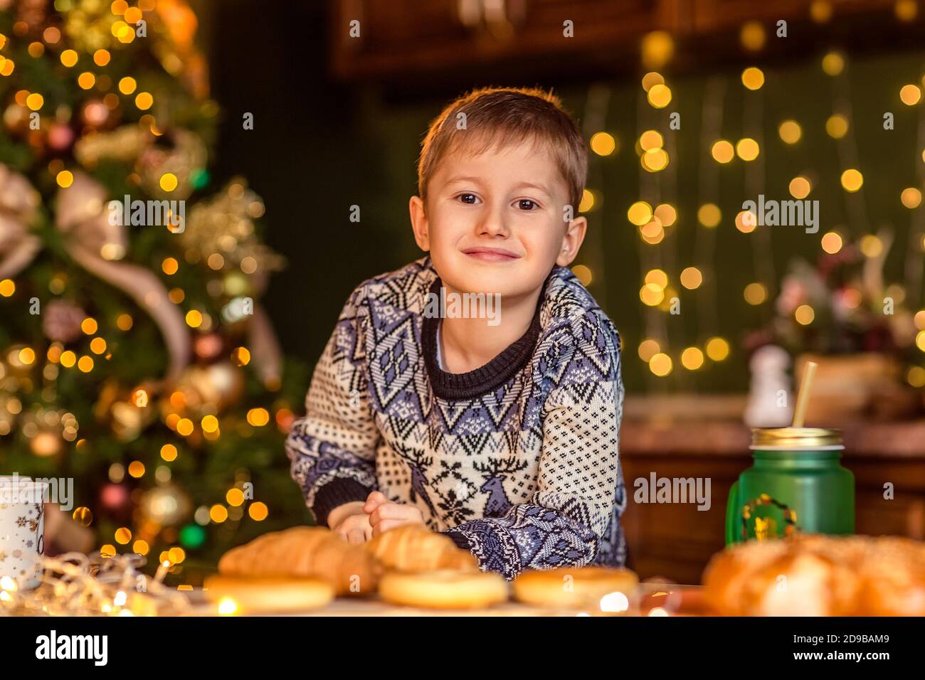 Un ragazzo si siede a un tavolo in una cucina di Natale decorata. Guarda il croissant e vuole mangiarlo. Accoglienti serate invernali a casa. Concetto di Cristo Foto Stock