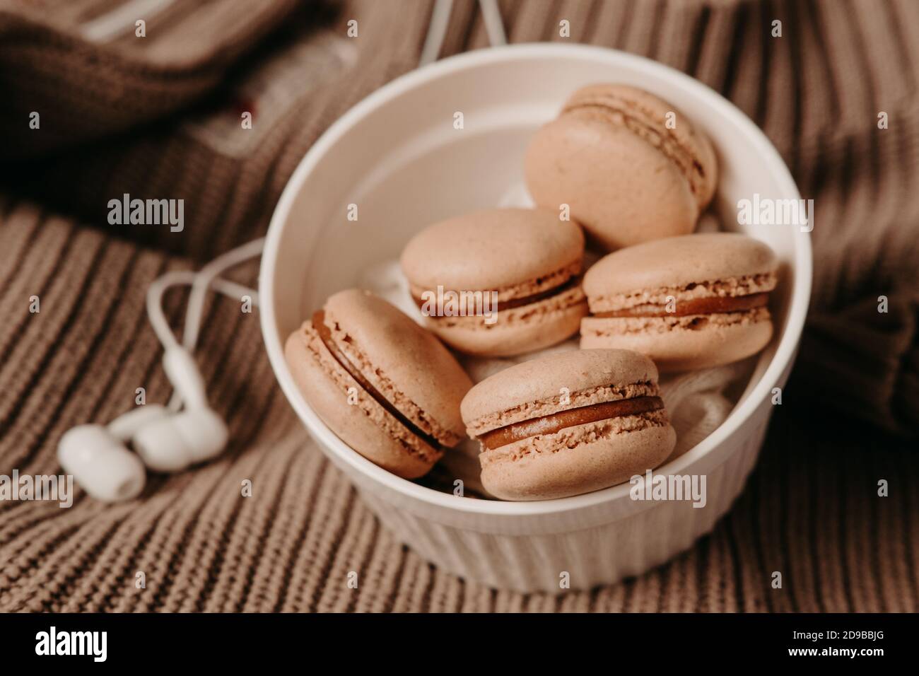 Cuffie bianche e macaroni al caramello su un maglione. Accogliente fotografia autunnale. Foto di cibo in tonalità marroni. Playlist invernale Foto Stock