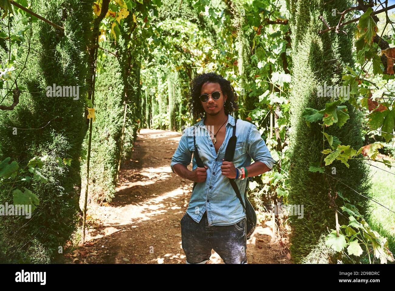 Musicista che cammina con la chitarra appesa alla schiena. Sta camminando in un parco con vegetazione. Foto Stock