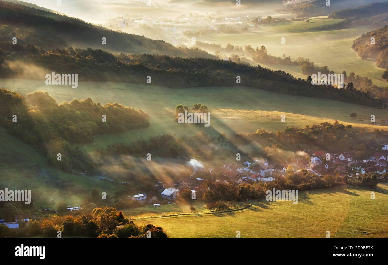 Vista aerea del villaggio in nebbia con raggi solari dorati all'alba in autunno. Bellissimo paesaggio rurale con strada, edifici, alberi colorati nebbia. Slovacco Foto Stock
