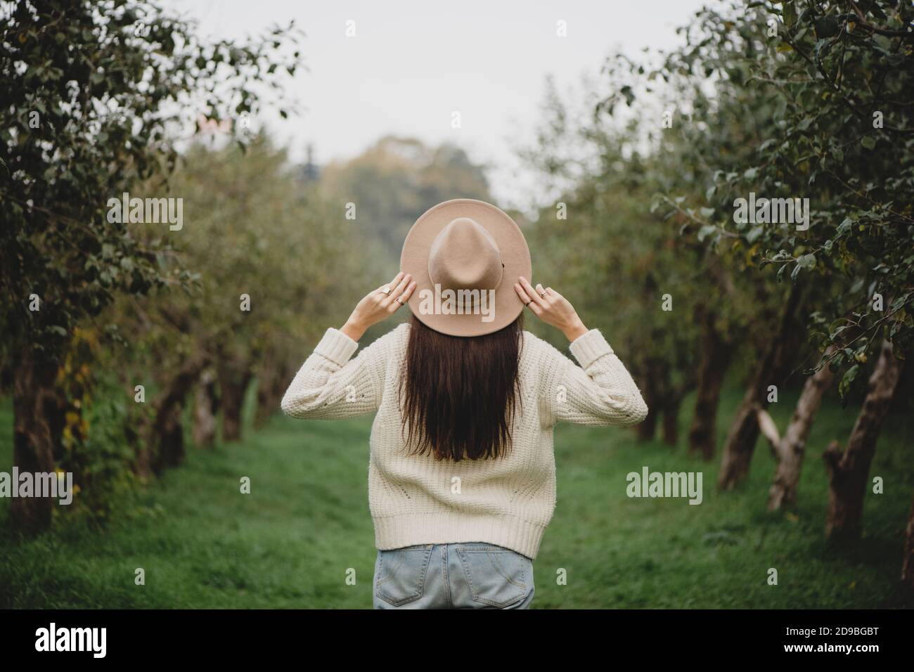 Vista posteriore di elegante donna dai capelli lunghi in cappello beige nel frutteto autunno. Foto Stock