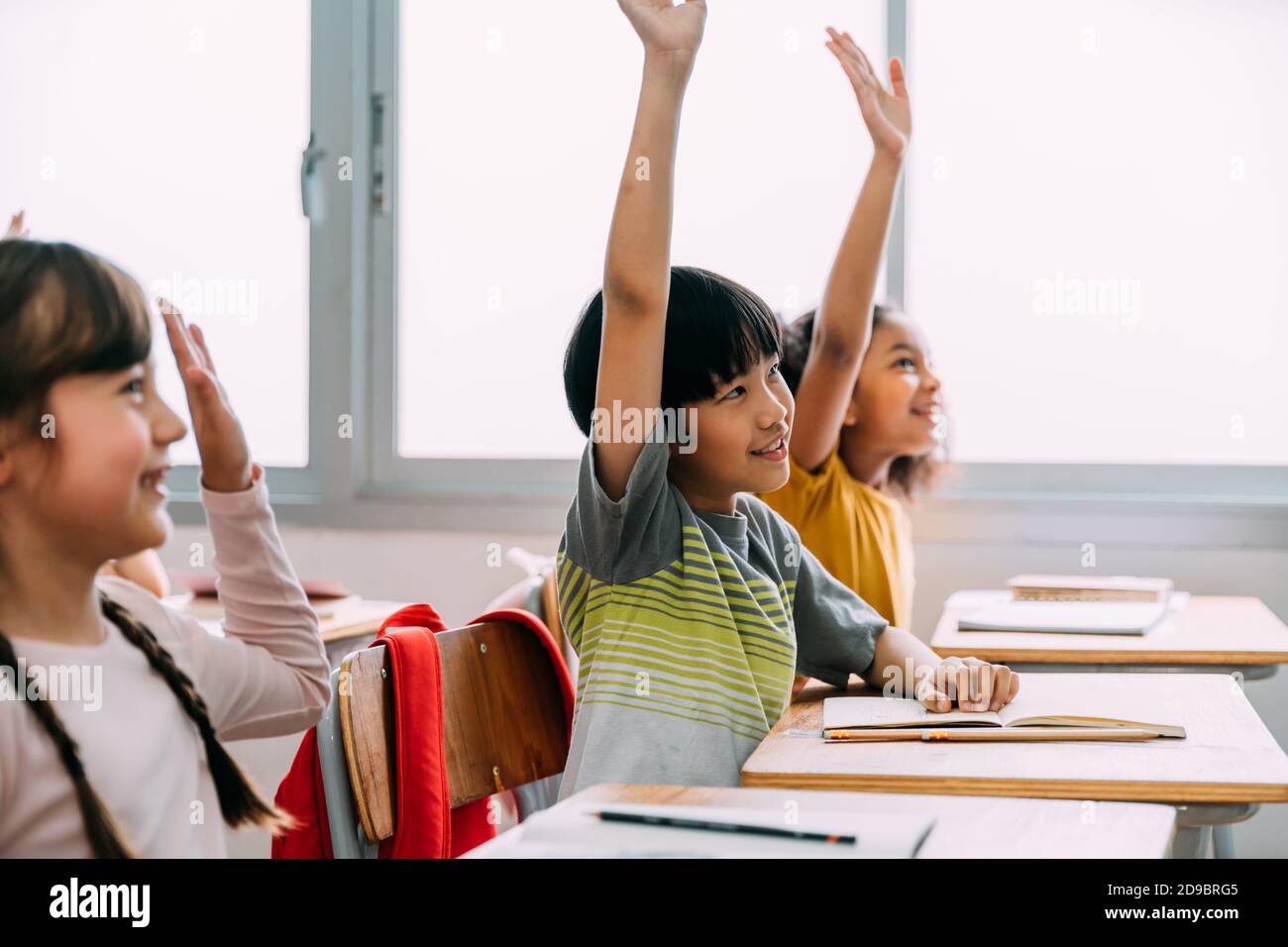 Il ragazzo asiatico di età elementare ha alzato le mani in su in Q ed IN UNA classe. Gruppo diversificato di alunni in età elementare nella scuola di costruzione dell'istruzione. Concetto di volontariato e partecipazione in classe. Foto Stock
