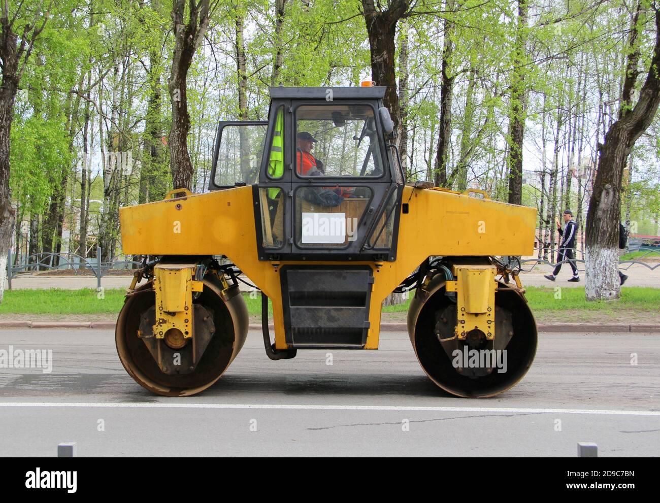 La pista di asfalto è gialla con ruote in ferro sulla strada in città sullo sfondo di alberi verdi. Lavori di riparazione su strada su asfalto di posa. Foto Stock