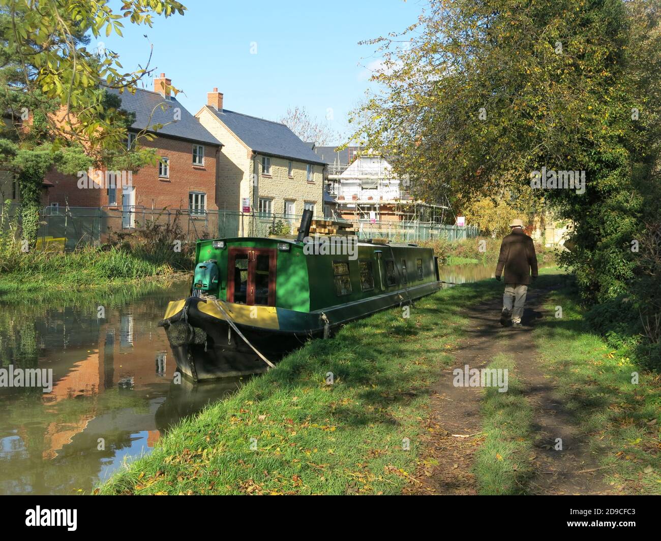 Un uomo cammina lungo l'alzaia passando un narrowboat che è ormeggiato di fronte alle nuove case che sono state costruite nel villaggio di Cosgrove, Northants. Foto Stock