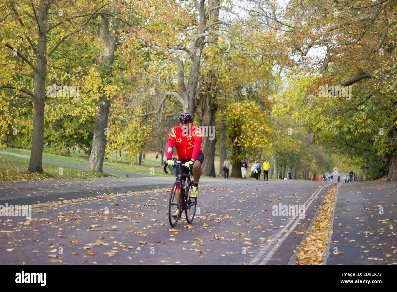 Acuto ciclista in cima rossa in salita strada coperta di foglie autunnali nel parco di Greenwich. Londra, Inghilterra, regno Unito Foto Stock