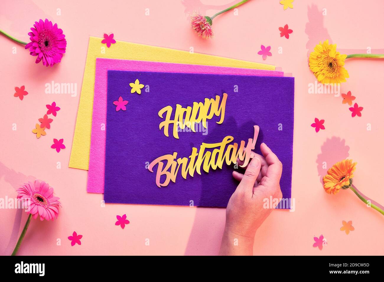 Vivace giacitura piatta con fiori di gerbera. Feltro viola con testo cartaceo buon compleanno in mano. Foto Stock