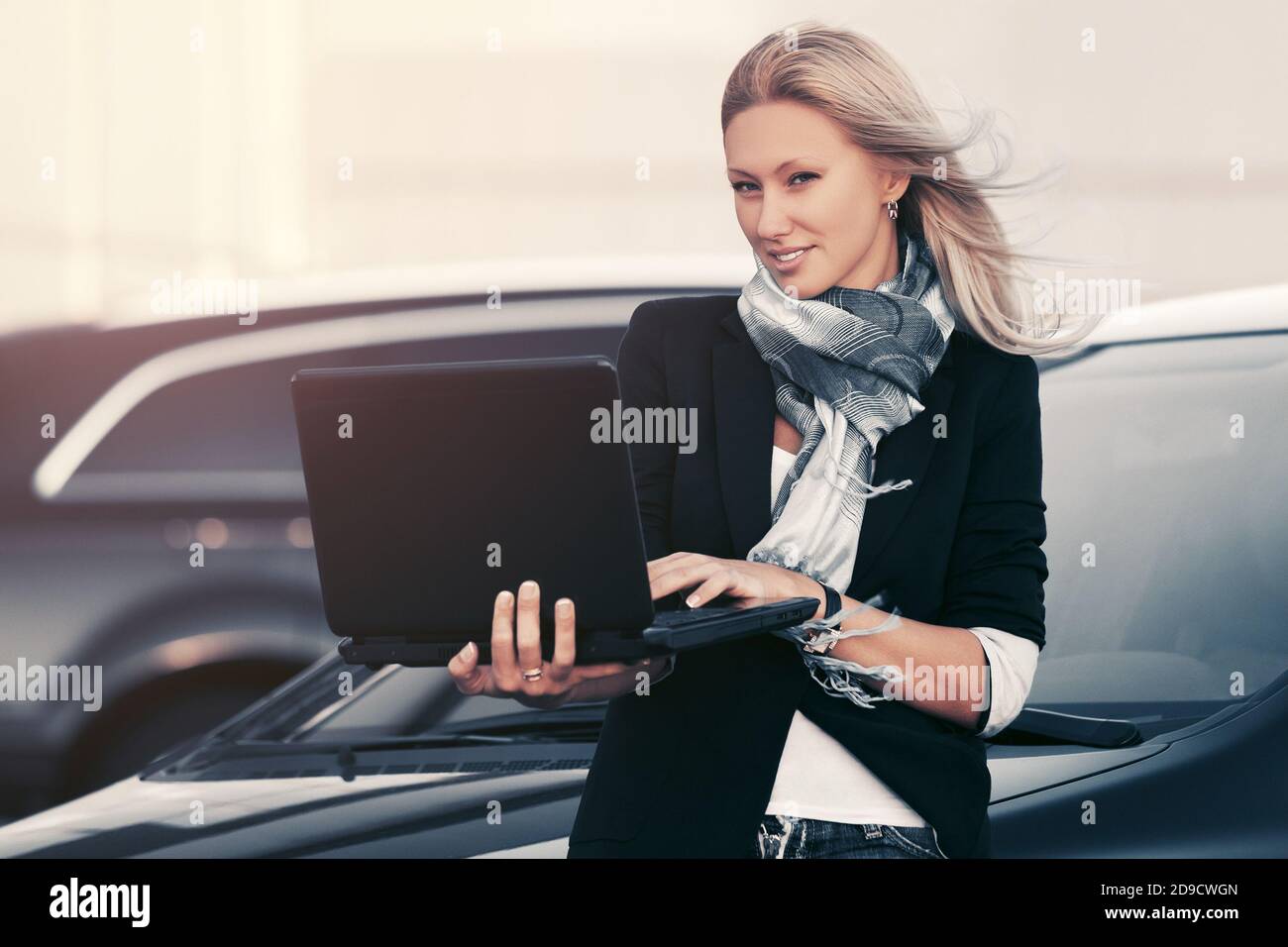 Giovane donna di affari di moda con il laptop appoggiato sulla sua auto Elegante modello femminile in blazer nero e sciarpa blu grigia Foto Stock