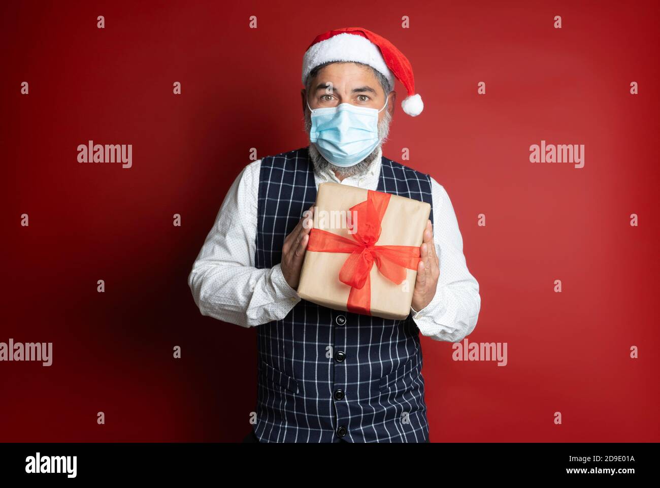 Uomo con maschera chirurgica e cappellino di natale tiene con illusione una confezione regalo su sfondo rosso Foto Stock
