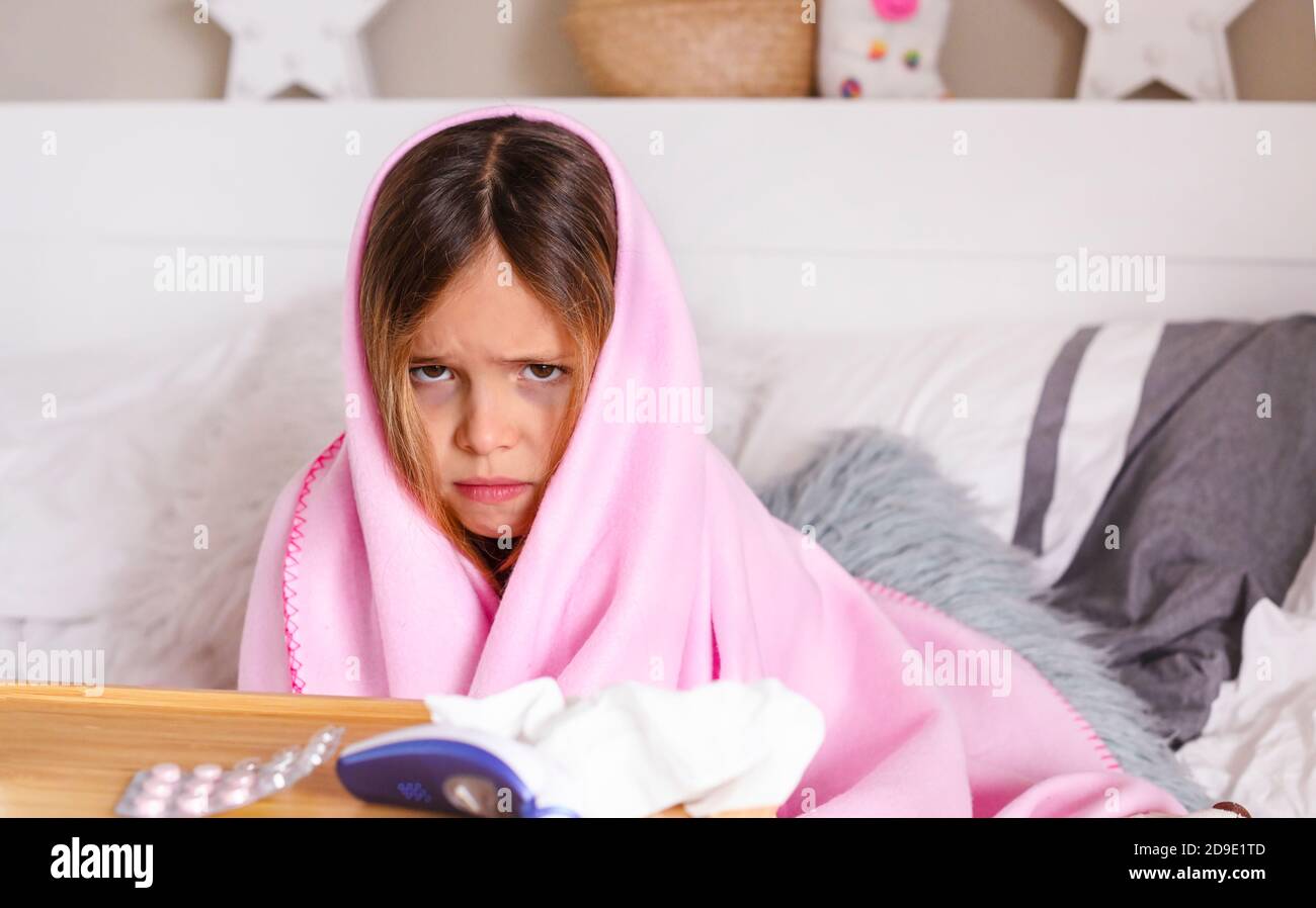 Una bambina dalle tristi emozioni, si avvolse in una coperta. Bambino con pillole e un termometro in camera da letto sul letto. Concetto di malattia virale e covidio 19. Foto Stock