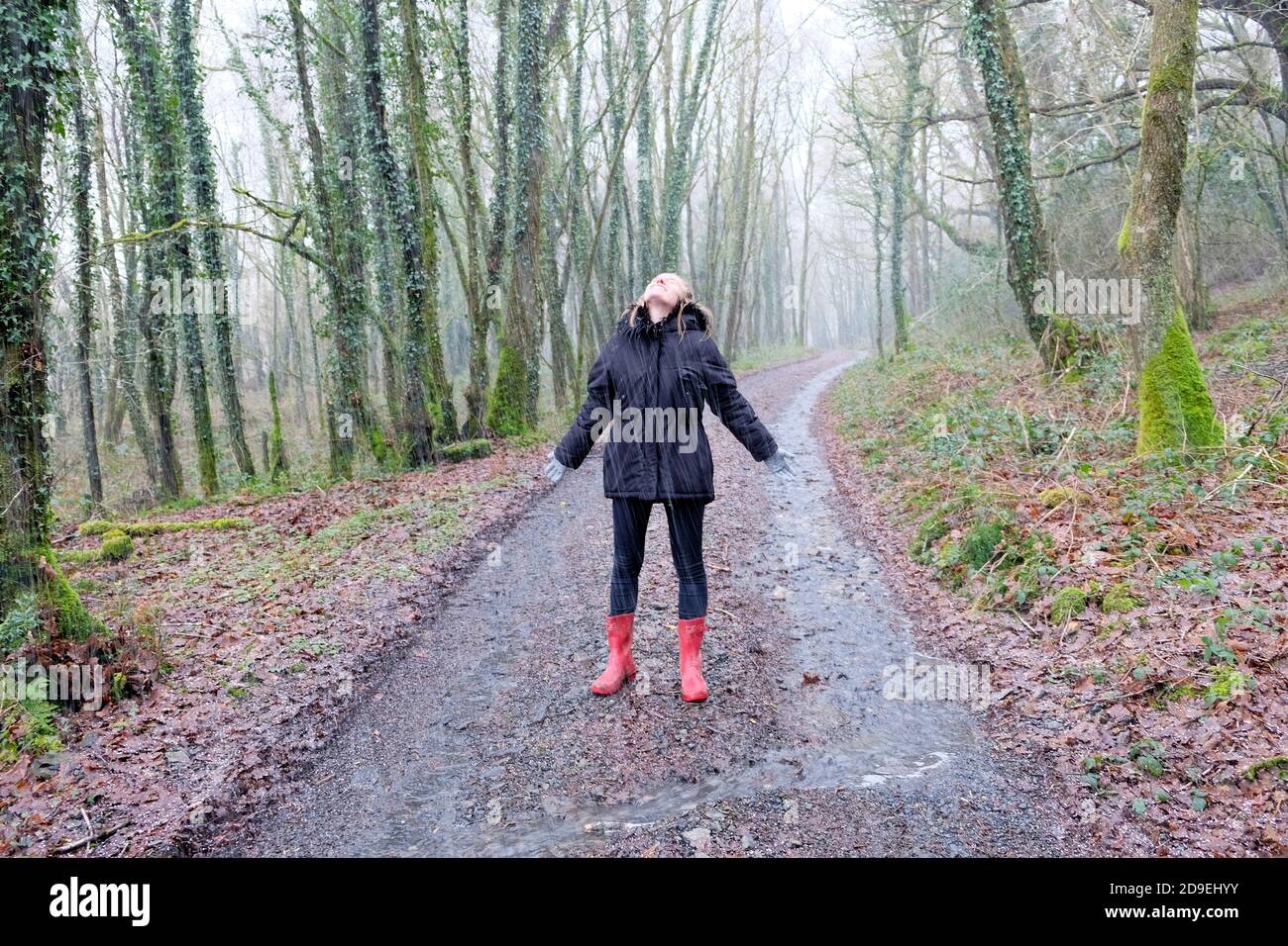 Giovane donna 20s indossando cappotto invernale wellies in piedi in pioggia giorno guardando in su con gioia raindrops cadere in faccia nella campagna Galles Regno Unito KATHY DEWITT Foto Stock