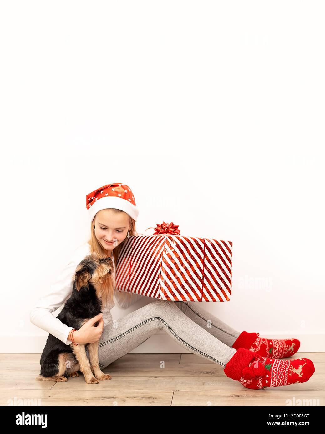 Una bella bambina in abiti casual con accessori di Natale si siede sul pavimento e abbraccia una grande scatola regalo con un nastro rosso e un arco. Lunghezza intera Foto Stock