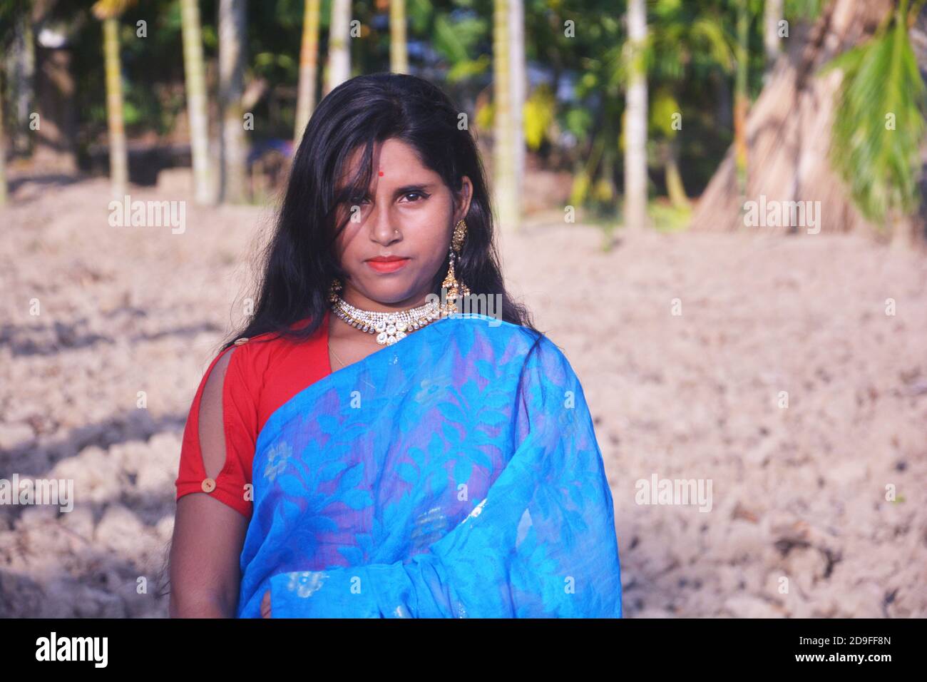 Primo piano di una ragazza indiana bengalese che indossa orecchini blu sari collana blusa rossa con lunghi capelli scuri in piedi in un campo arato. Foto Stock