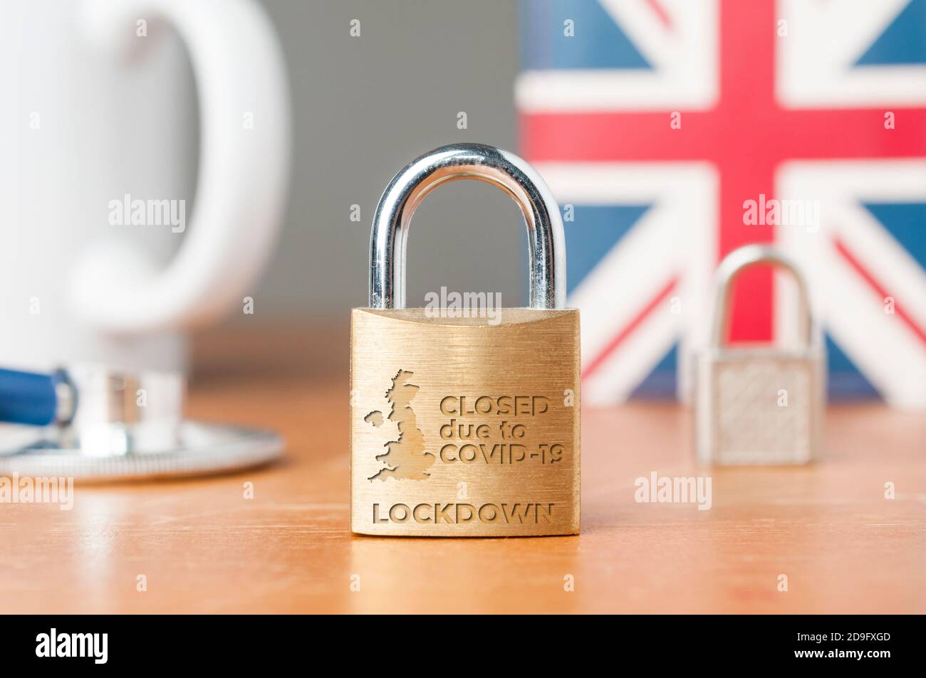 Concetto di blocco Covid-19 UK. Una serratura con il messaggio 'chiudere a causa di covid-19 blocco' su un tavolo di legno con Union Jack sullo sfondo, una tazza e. Foto Stock