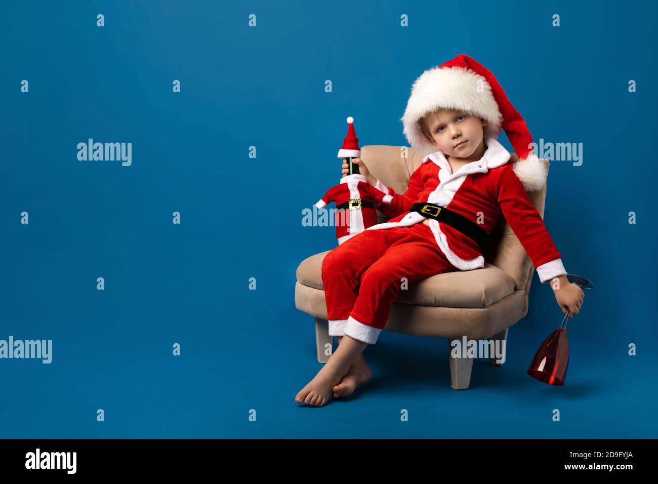 Carismatico bambino vestito da Babbo Natale con una bottiglia di champagne  e un bicchiere vuoto su sfondo blu, guardando la macchina fotografica.  Natale e N Foto stock - Alamy
