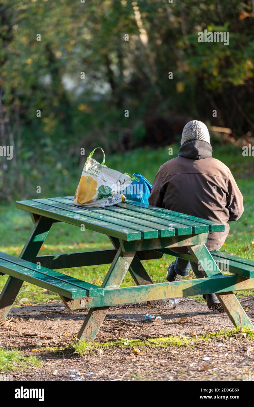 un uomo che indossa un cappello seduto da solo a una panca di parco e tavolo con le sue borse di spesa e di trasporto accanto a lui sul tavolo. Foto Stock