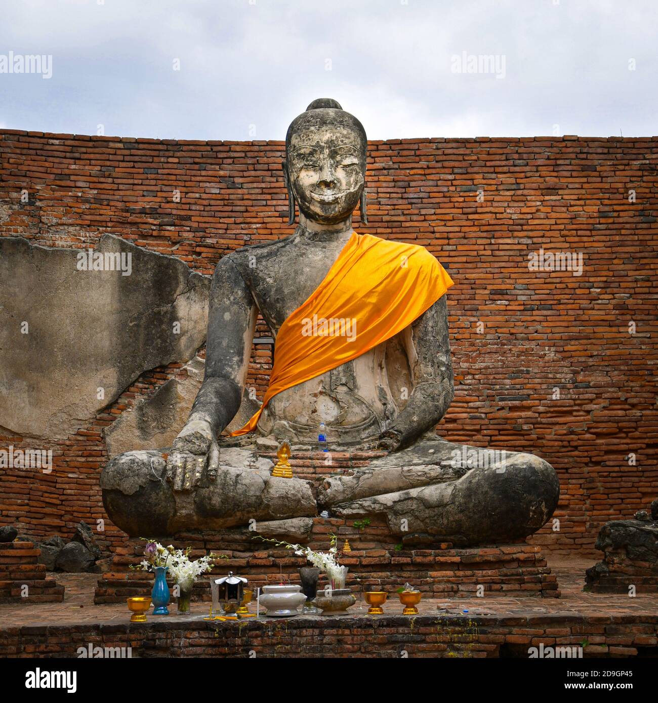 Una statua del buddha seduto che indossa un telaio di zafferano in un cortile di mattoni di un tempio buddista tailandese. Di fronte alla statua si trovano una varietà di offerte Foto Stock