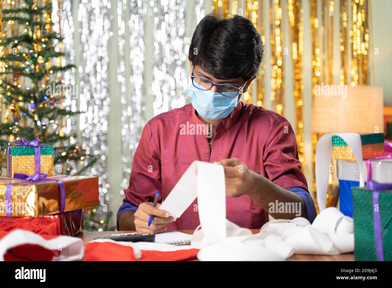 Giovane uomo in maschera medica occupato nel calcolo delle spese di vacanza fatture con sfondo decorato natale e regali davanti al tavolo. Foto Stock