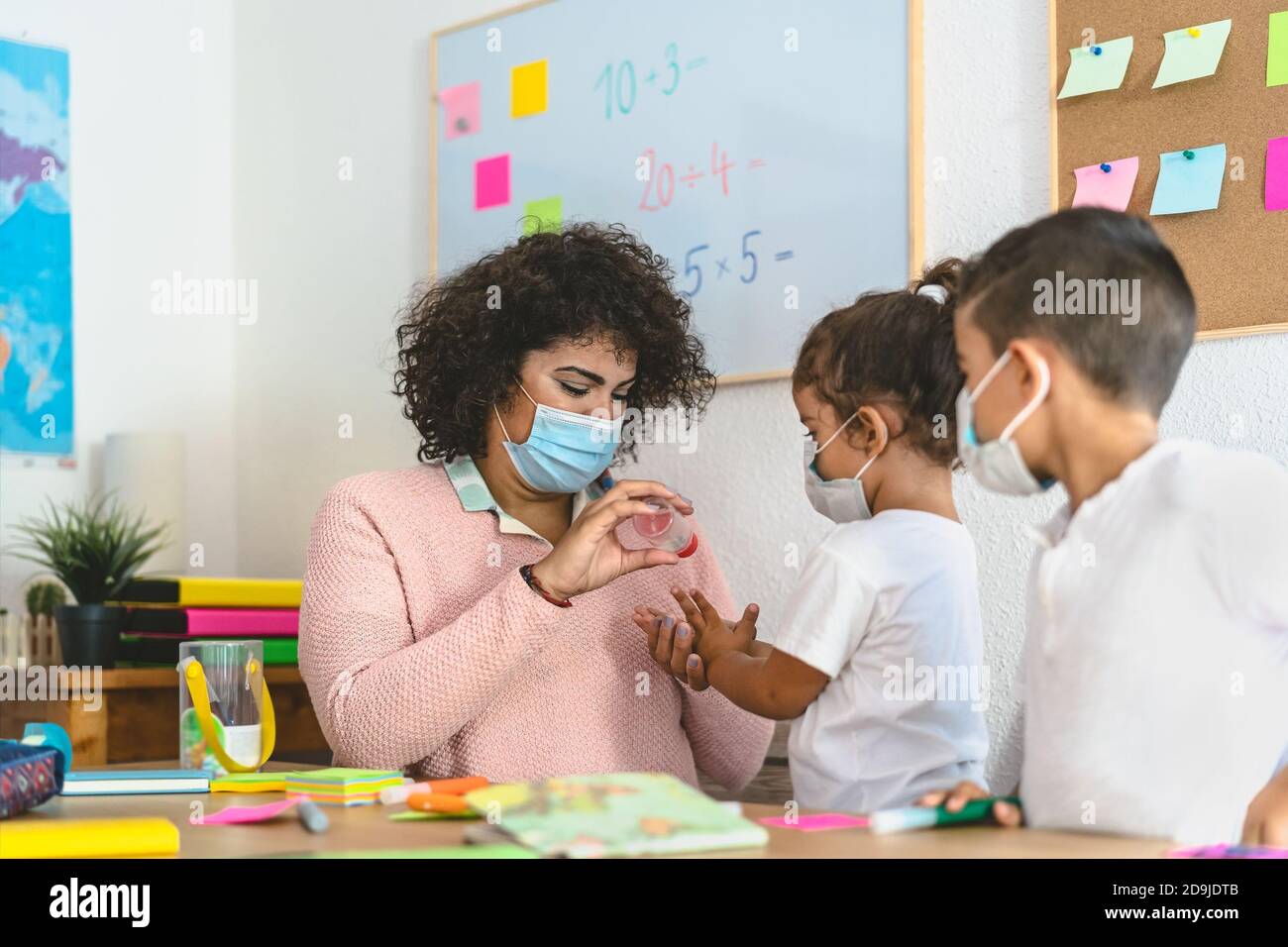 Insegnante di pulizia mani ai bambini studenti con gel igienizzante mentre indossare la maschera facciale in classe prescolare durante la pandemia del virus corona Foto Stock