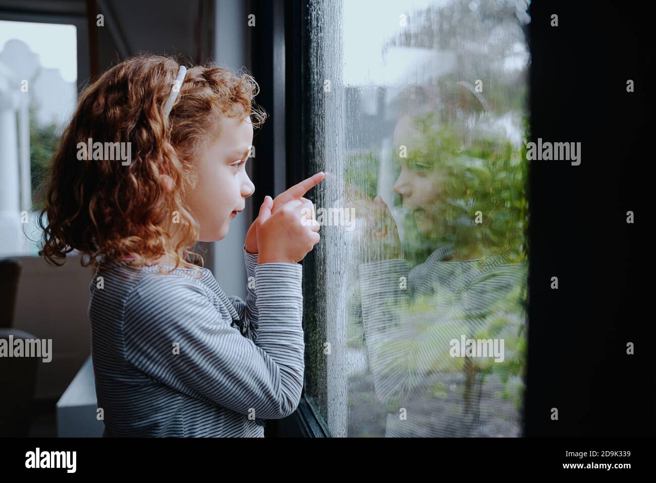 Carina bambina in piedi vicino alla finestra guardando la pioggia all'esterno Foto Stock