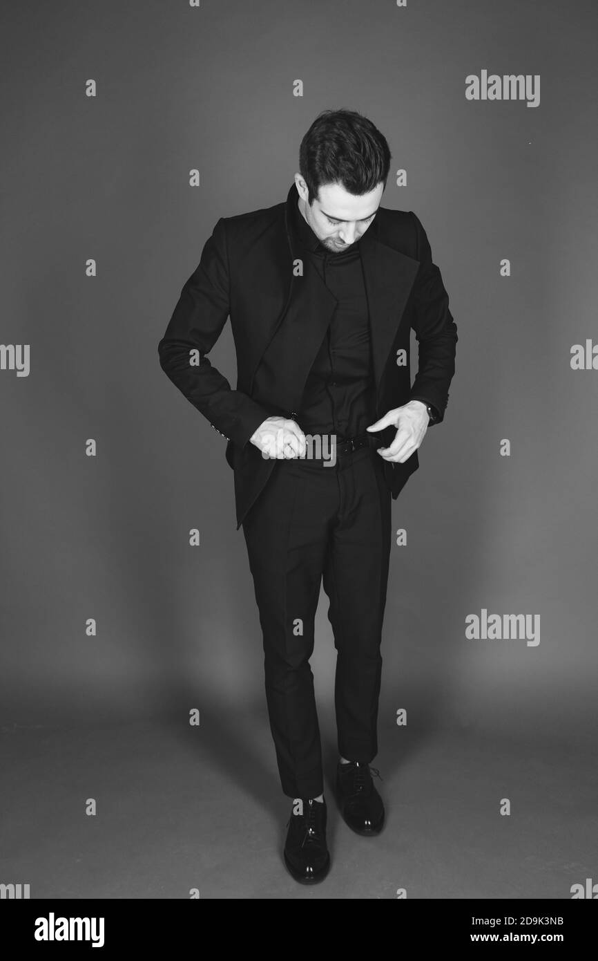 Studio ritratto di un giovane caucasico in un blazer nero, guardando verso il basso, calzando la cintura, in piedi su uno sfondo di studio semplice Foto Stock