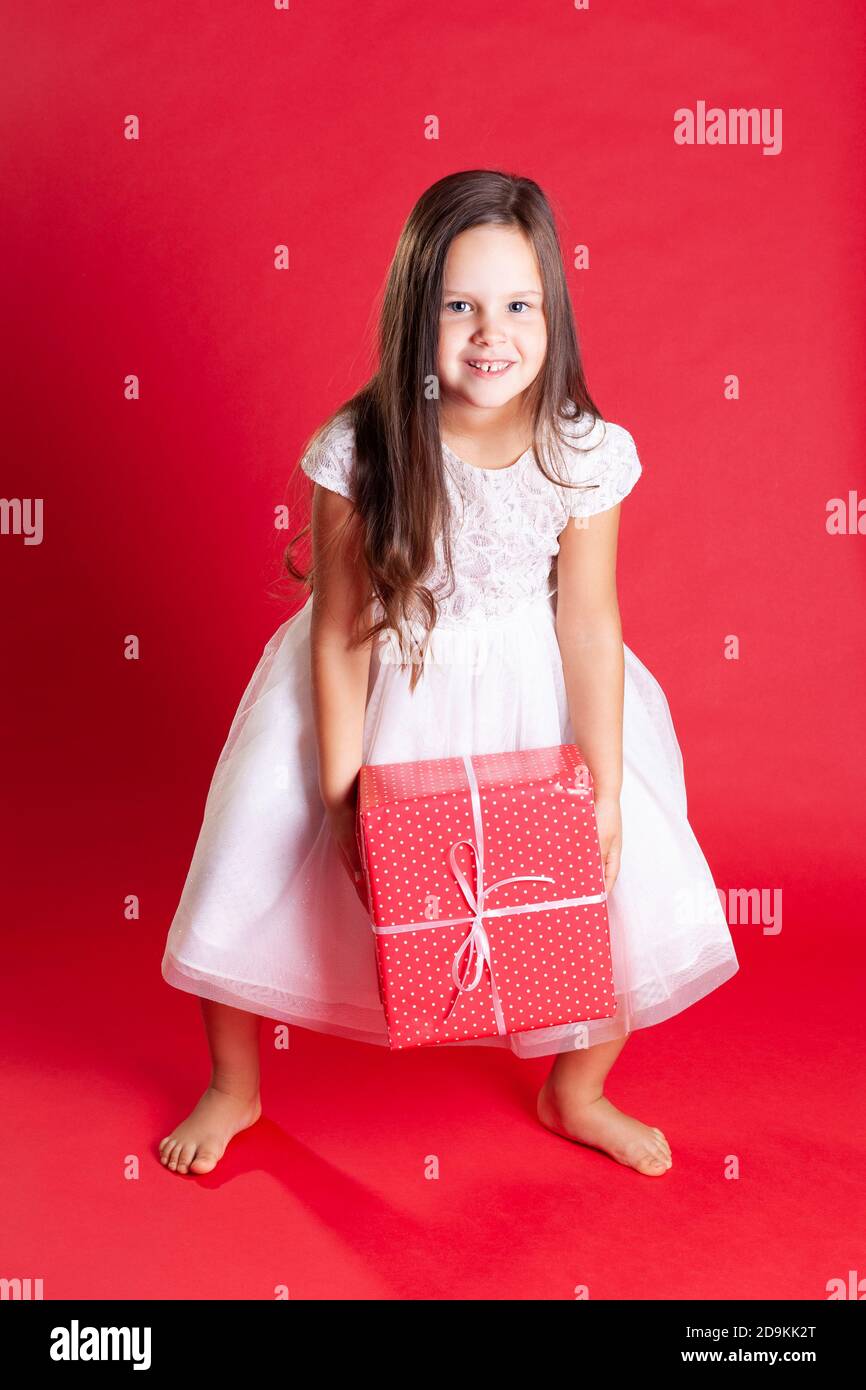 bella bambina brunetta in abito bianco pick up confezione regalo Dal pavimento per Natale o compleanno su sfondo rosso isolato Foto Stock