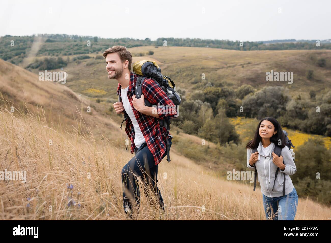 Uomo sorridente con zaino che cammina sulla collina vicino afroamericano ragazza durante la vacanza Foto Stock