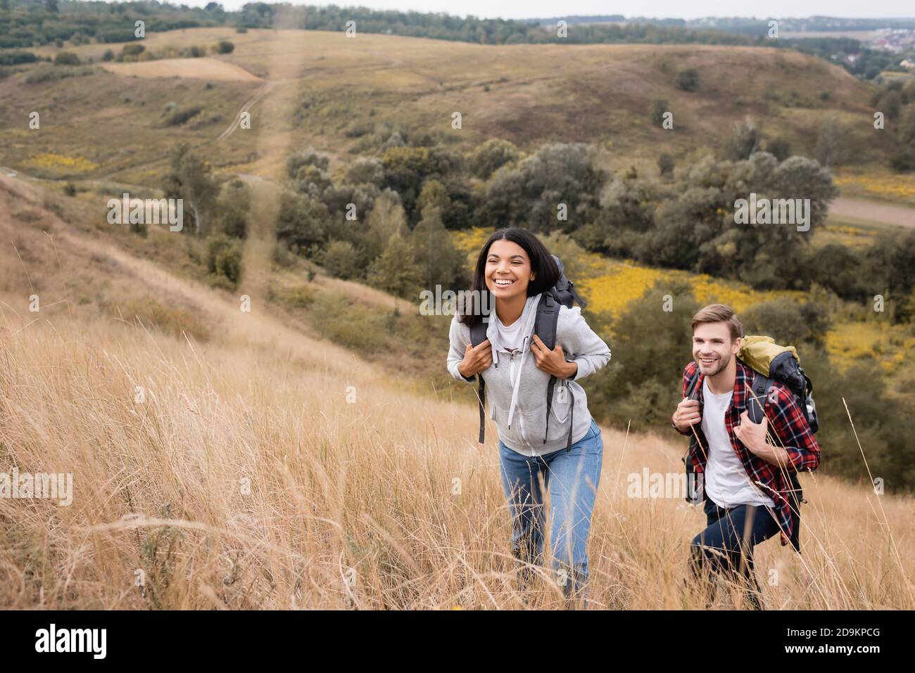 Coppia interracial con zaini che camminano su collina con erba durante viaggio Foto Stock