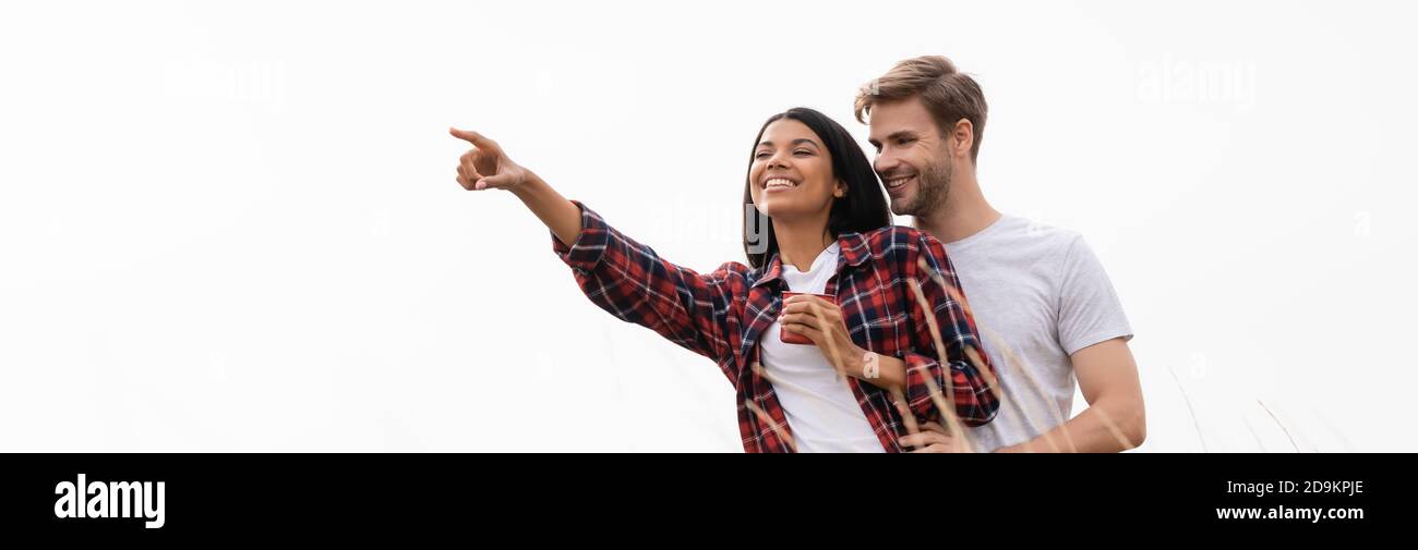 Uomo sorridente abbracciando la ragazza afroamericana con tazza vicino erba isolata su bianco, bandiera Foto Stock