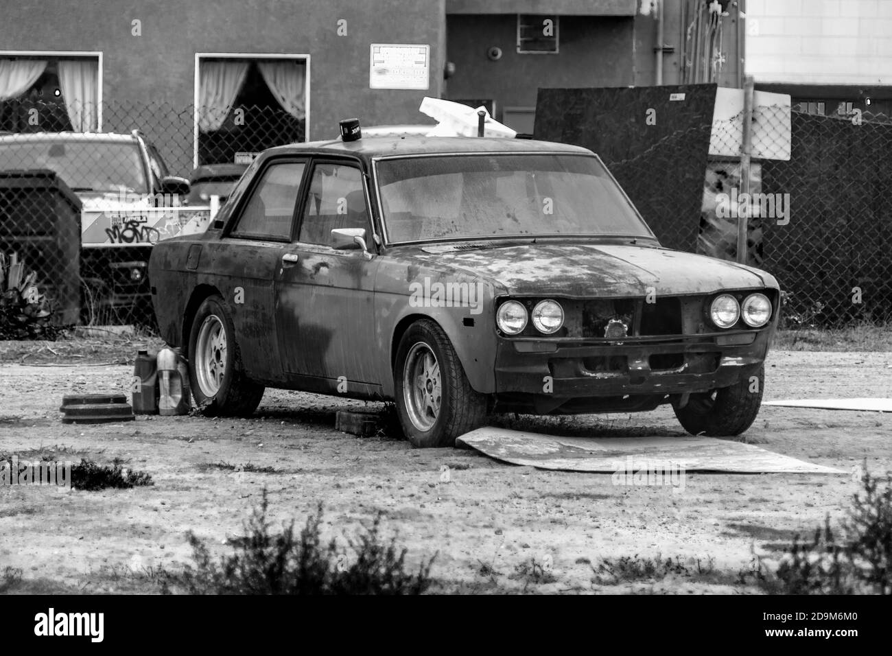 Immagine in bianco e nero di un'auto arrugginita, vecchia e classica in un lotto vuoto di sporcizia pieno di erbacce. Foto Stock