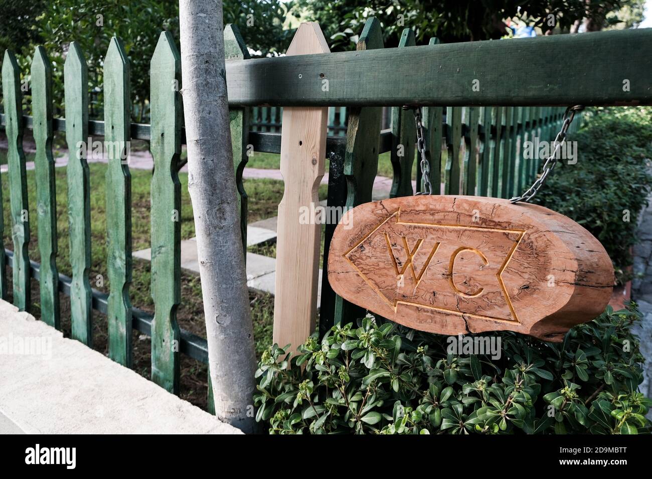 Tavola di legno marrone con segno di WC intagliato e freccia per mostrare la direzione ad un bagno pubblico. Bella segnaletica vicino recinzione verde all'aperto. Foto Stock