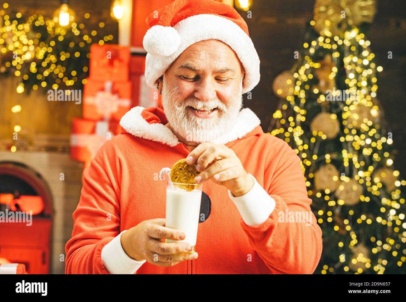 Santa tenuta cookie e bicchiere di latte sullo sfondo dell'albero di Natale. Babbo Natale con biscotti natalizi e latte sullo sfondo dell'albero di Natale. Foto Stock