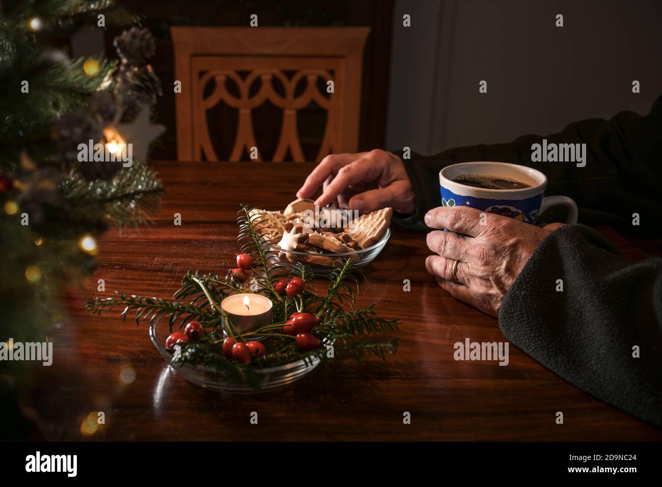 Mani di un uomo anziano seduto da solo ad un tavolo con biscotti di Natale, caffè e decorazione festosa accanto ad una sedia vuota, vacanze solitarie Foto Stock
