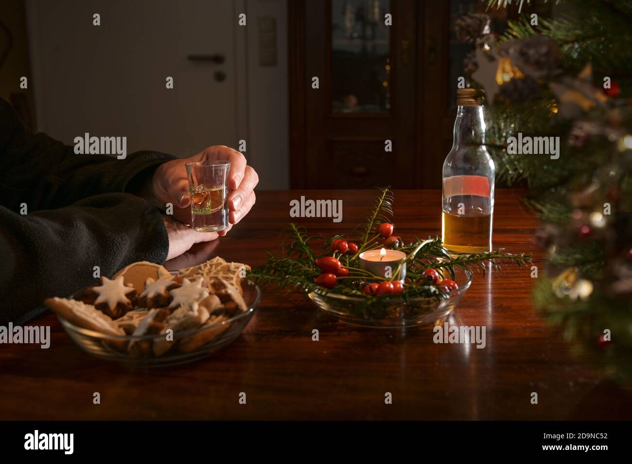 Mani di un uomo anziano che tiene un vetro dello shot da solo ad un tavolo con decorazione di Natale, biscotti, candela e una bottiglia mezzo-vuota di liquore, solitario hol Foto Stock