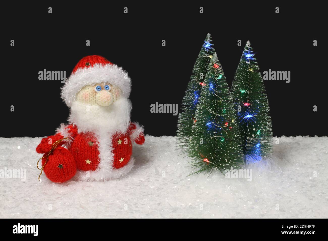 Il giocattolo in lana maglia Babbo Natale si trova accanto al giocattolo verde alberi di Natale con luci sulla neve bianca artificiale su sfondo nero. Primo piano Foto Stock