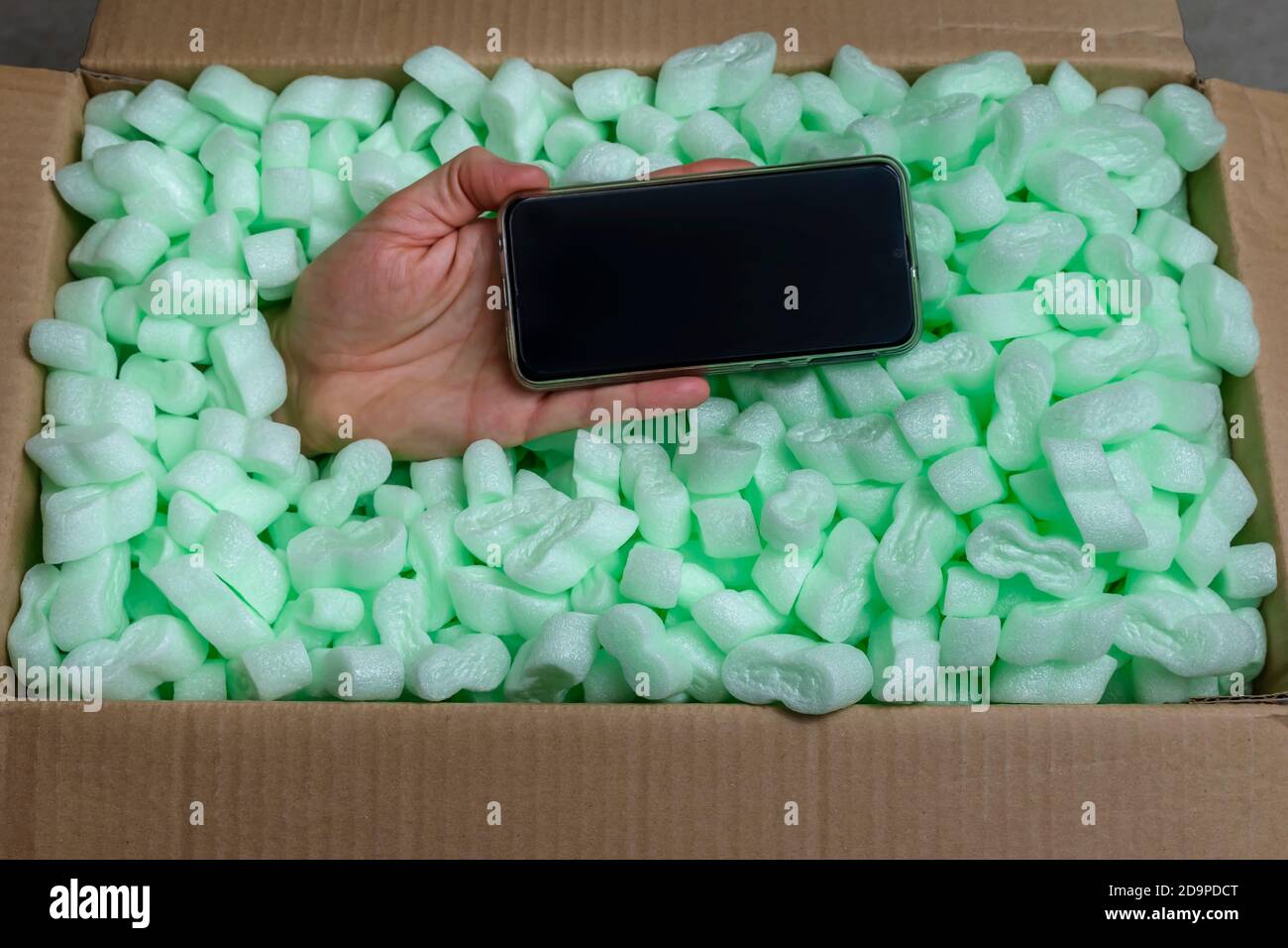 Una mano che tiene uno smartphone emerge da una scatola di cartone piena di trucioli di polistirene Foto Stock
