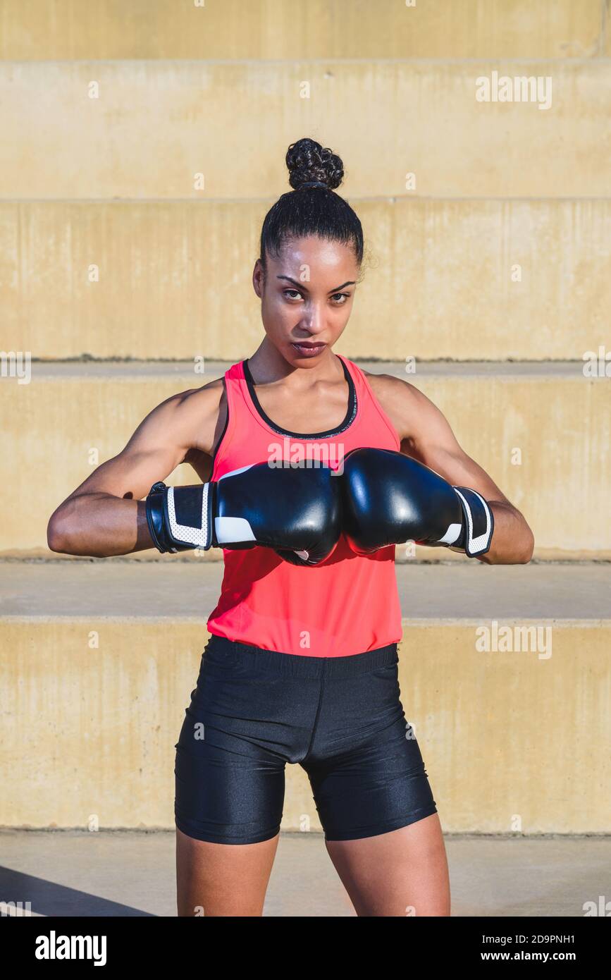 ritratto verticale di giovane donna afro americana attraente e muscolare indossare abbigliamento sportivo nero e rosa guardando la fotocamera e urti guanti da boxe b Foto Stock