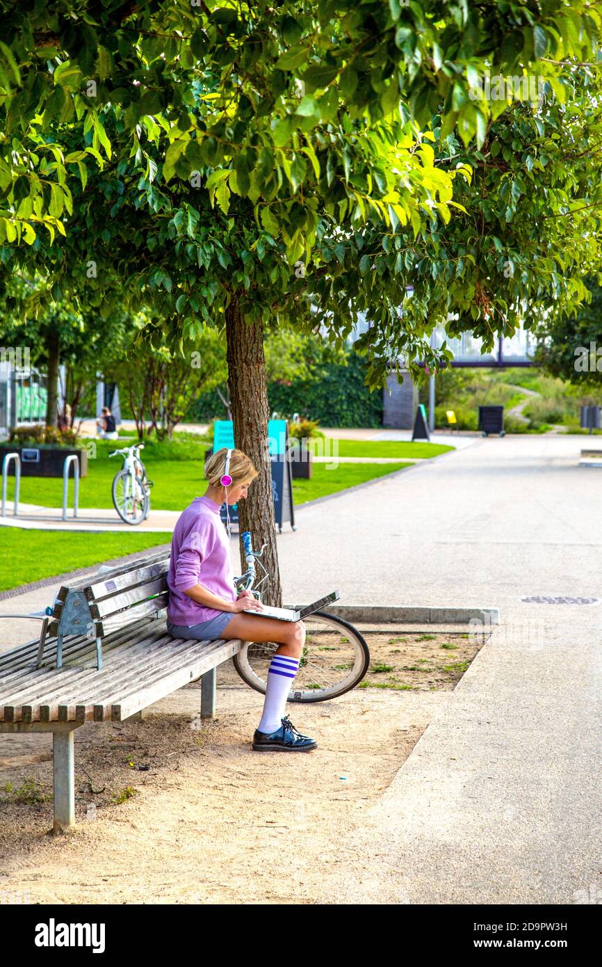 Giovane donna seduta su una panchina, cuffie, ascolto di musica e lavoro su un computer portatile con la sua bicicletta sullo sfondo, Stratford, Londra, Regno Unito Foto Stock
