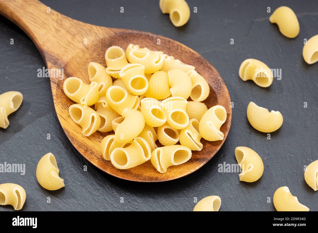 I gomiti crudi pasta macaroni in cucchiaio di legno su tavola scura. Foto Stock