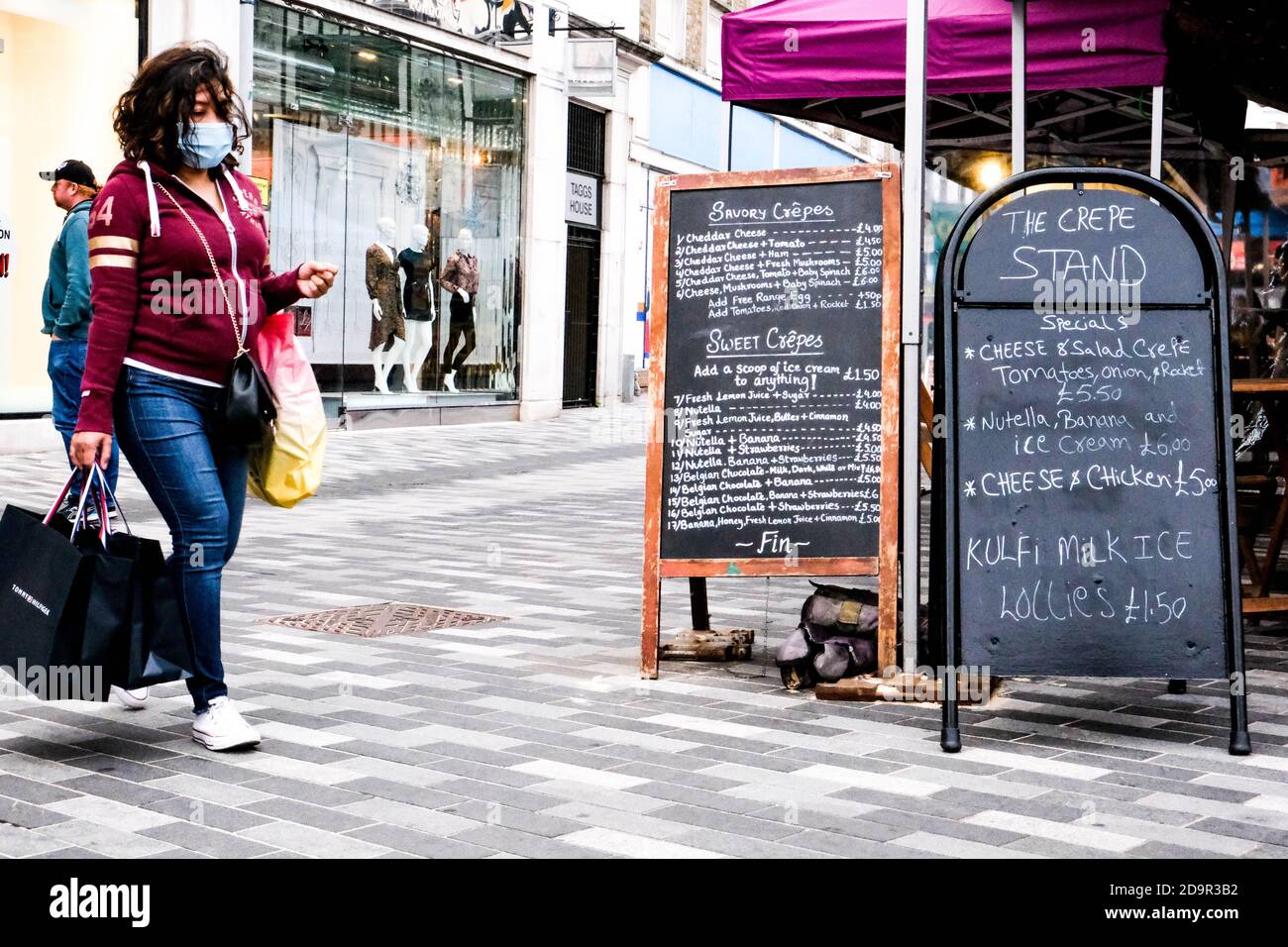 Donna Nera Passeggiate all'esterno DI un negozio durante il COVID-19 Coronavirus indossare Rivestimenti facciali di protezione e trasporto DI una borsa Foto Stock