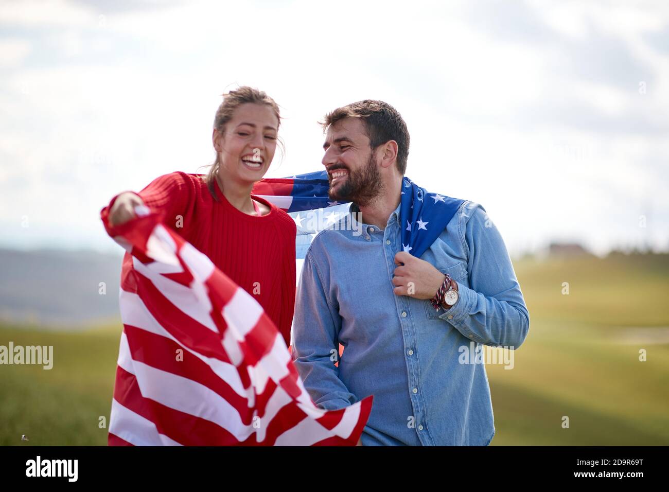 Una coppia allegra e allegra vestita di bandiera americana in un prato in una bella giornata di sole. Concetto di elezione, campagna, libertà Foto Stock