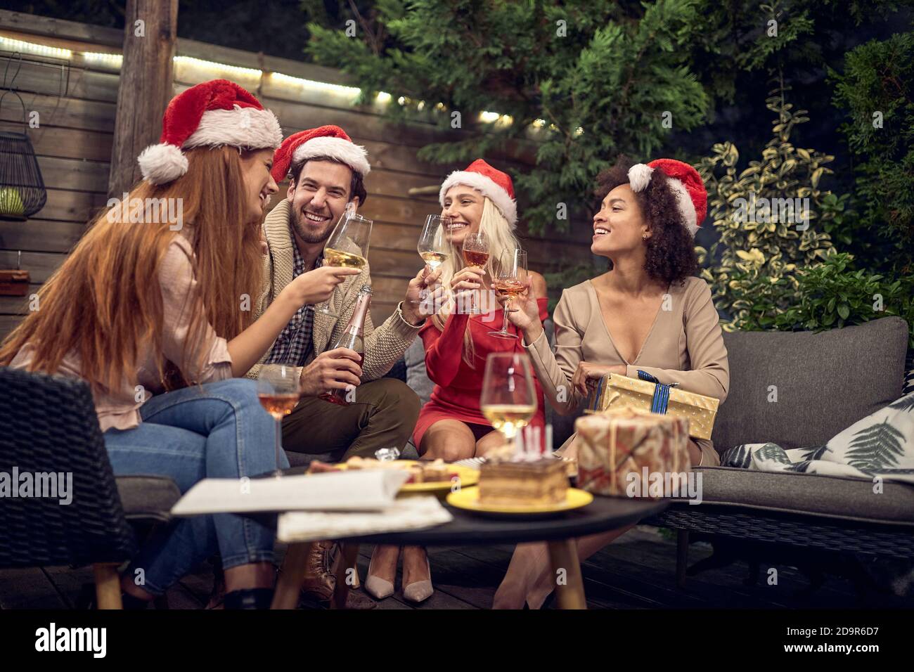 gruppo di amici che brindano con bicchieri da vino, festeggiano il nuovo anno, indossando cappelli santa clausola, seduti all'aperto nel cortile Foto Stock