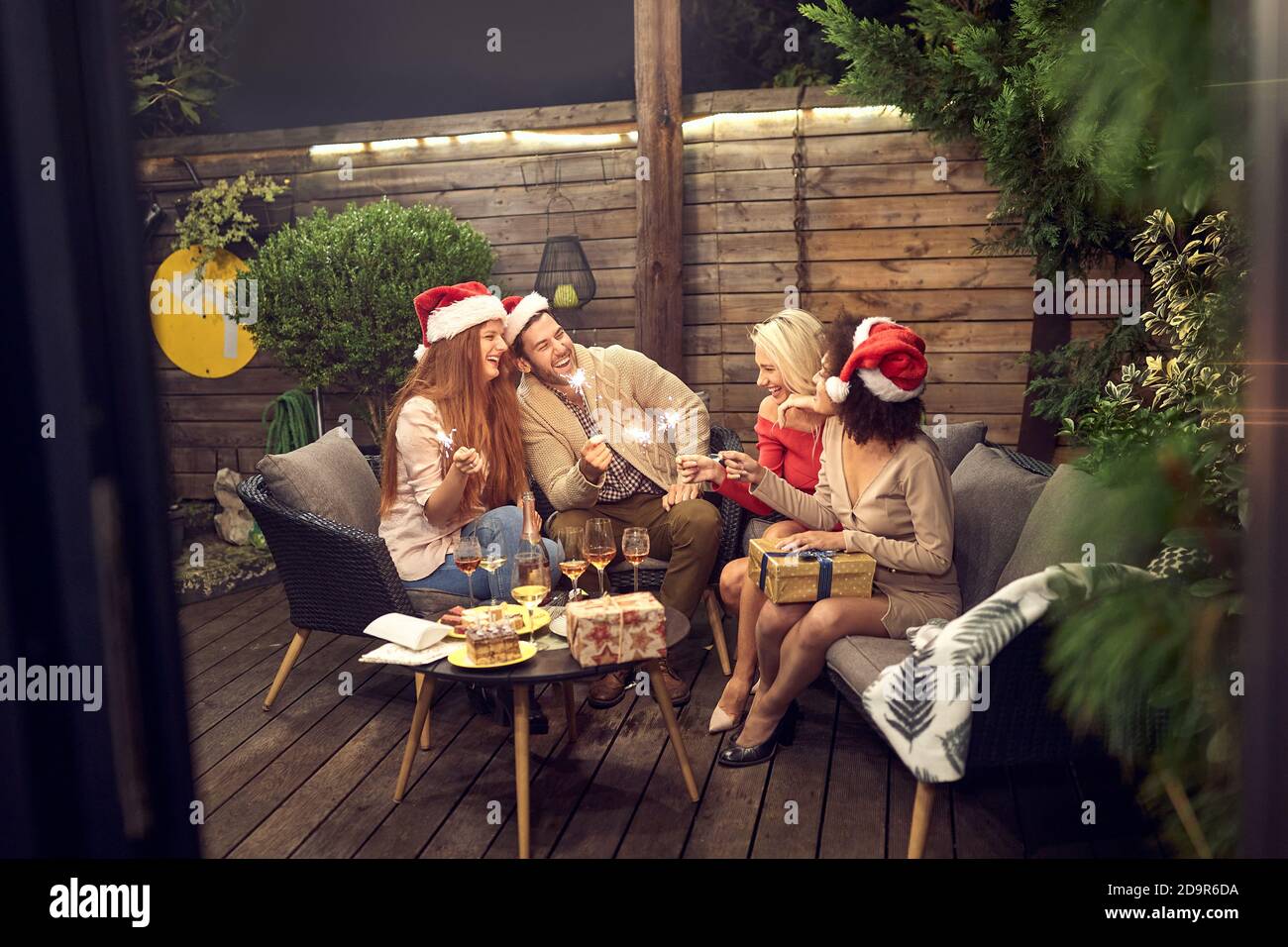 gruppo di giovani amici adulti che festeggiano il nuovo anno all'aperto, seduti nel cortile con frizzanti bastoni in mano indossando cappelli santa Foto Stock