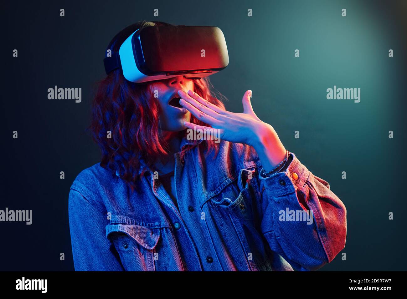 Espressione facciale di giovane ragazza con occhiali di realtà virtuale entra in un neon rosso e blu in studio Foto Stock
