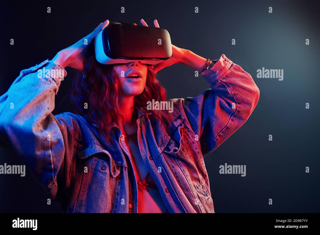 Espressione facciale di giovane ragazza con occhiali di realtà virtuale entra in un neon rosso e blu in studio Foto Stock
